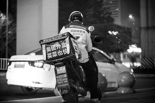 Black and white photo of a delivery driver on a scooter in urban city streets at night.