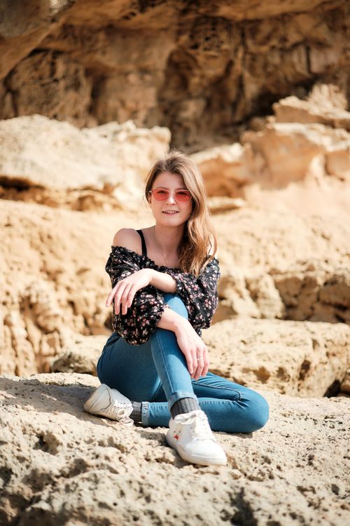 Free A fashionable young adult woman with sunglasses and jeans sitting and smiling on rocky terrain in Ayia Napa, Cyprus. Stock Photo