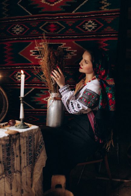 A woman in traditional Ukrainian attire sits by candlelight, holding dried plants.