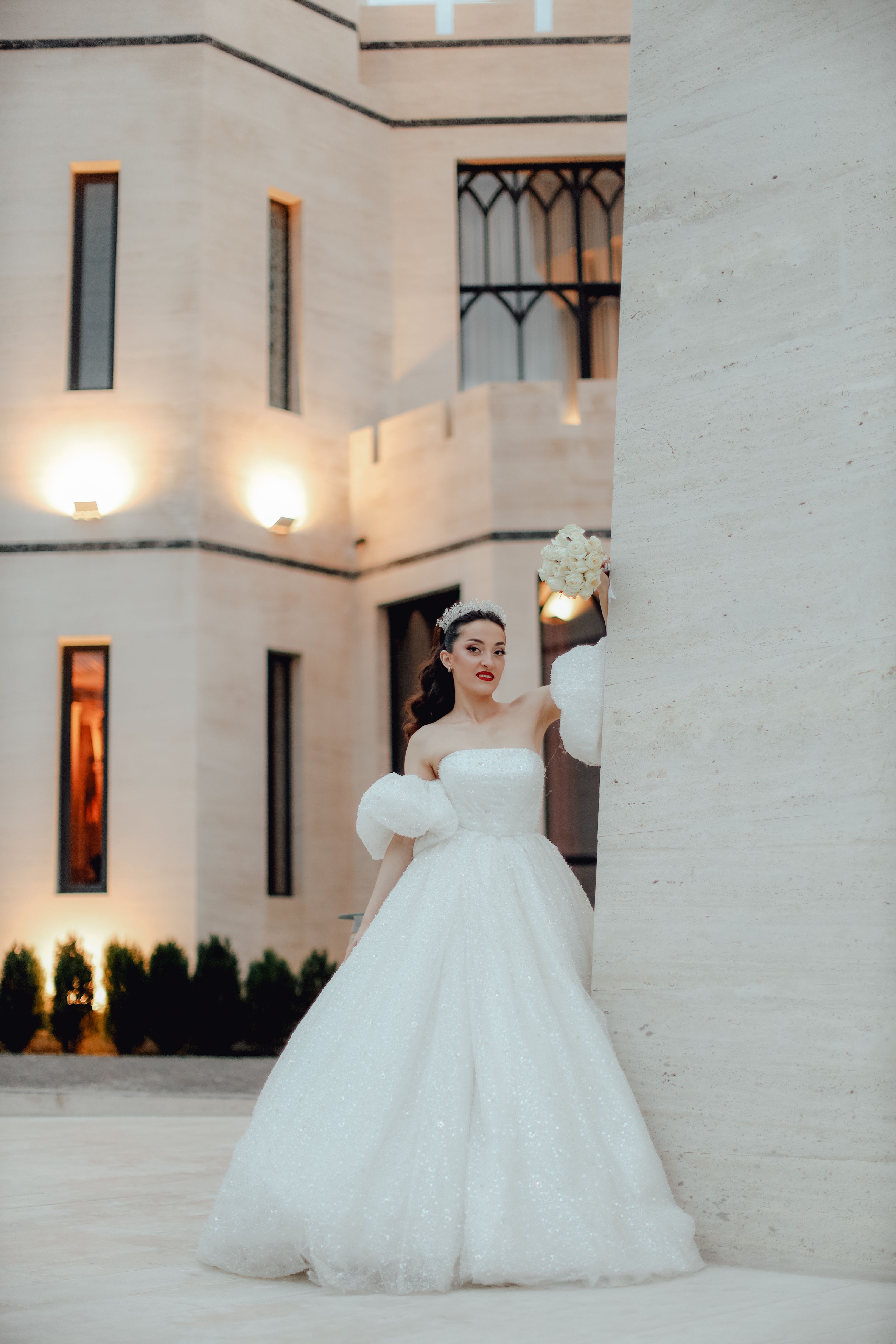 A stunning bride in a white wedding dress stands elegantly by a modern building holding a bouquet.
