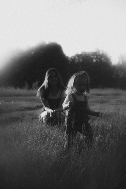 Black and White Photo of a Girl Looking at a Toddler Run on a Meadow 