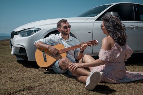 Woman in Dress Sitting with Man Playing Guitar by Car