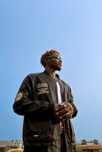 Confident young man in sunglasses with locs and rings, standing under a clear sky.
