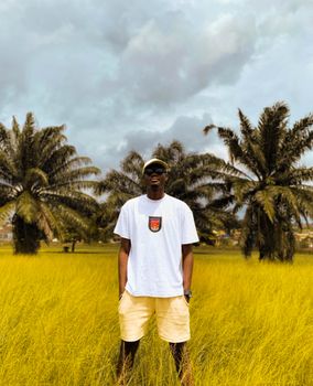 A young man stands in a tropical field with palm trees in Ibadan, Nigeria, wearing casual summer attire.