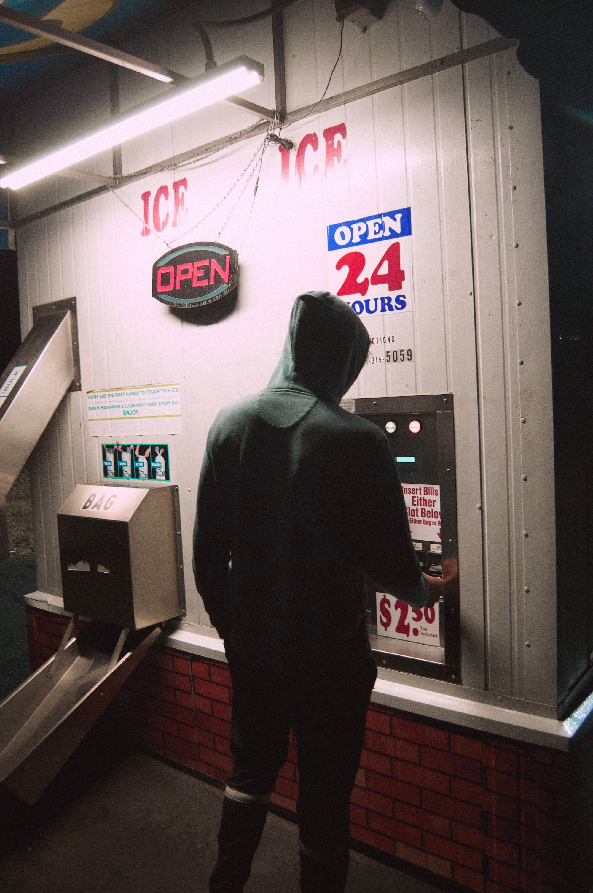A man in a hoodie uses an ice vending machine on a dimly lit street corner at night.
