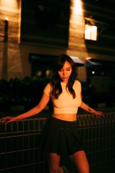 A young woman posing by a fence in an urban setting under warm, illuminated lights at night.