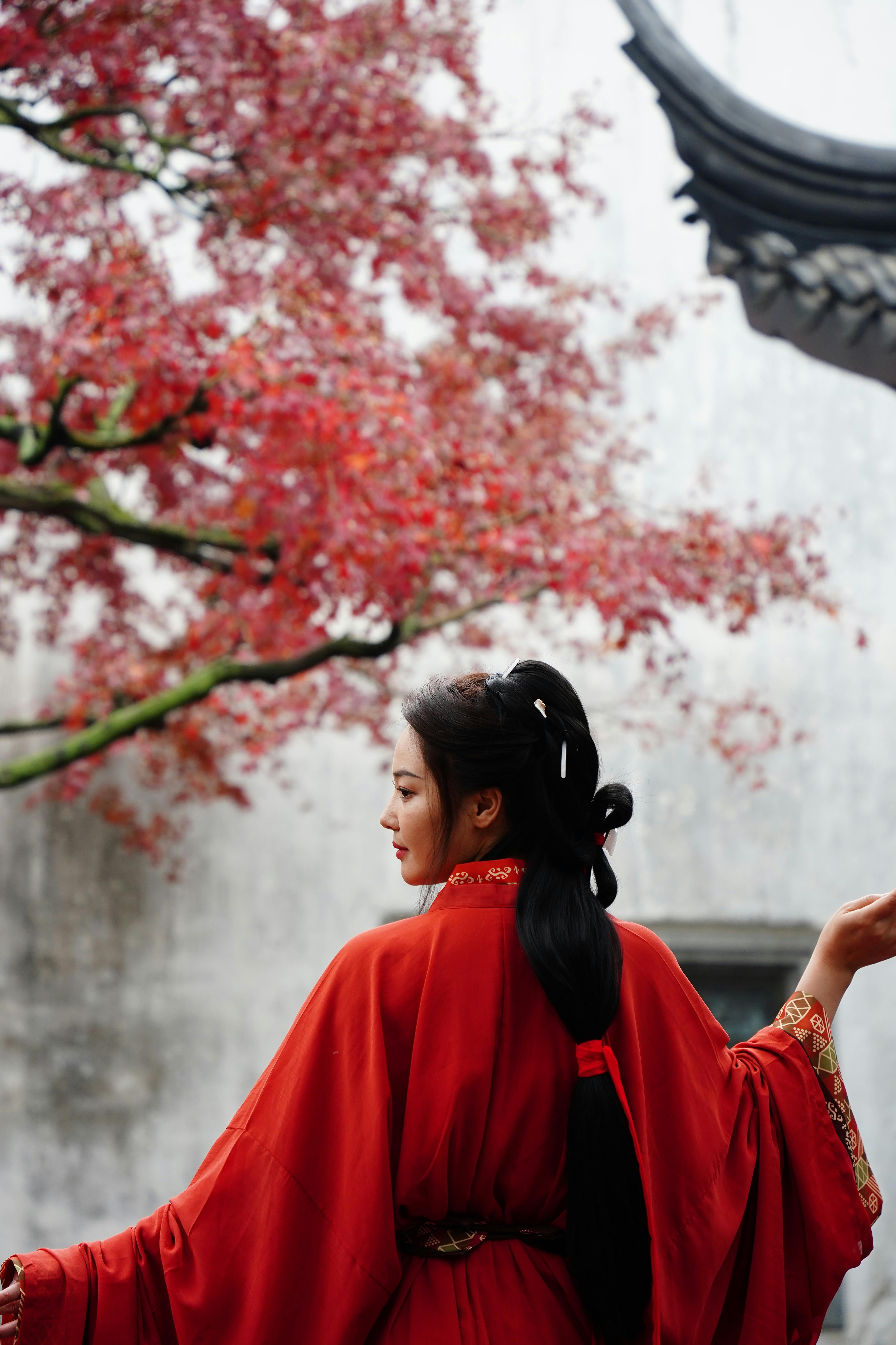 A woman in a red dress stands elegantly, with cherry blossoms in the background, capturing traditional beauty.
