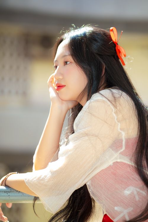Free A young Asian woman stands outdoors, bathed in sunlight, gazing peacefully with a red bow in her hair. Stock Photo