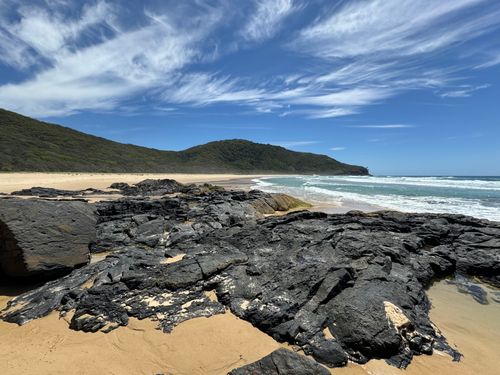 Free A rugged beach with black rocks and waves under a bright blue sky, ideal for summer travel. Stock Photo