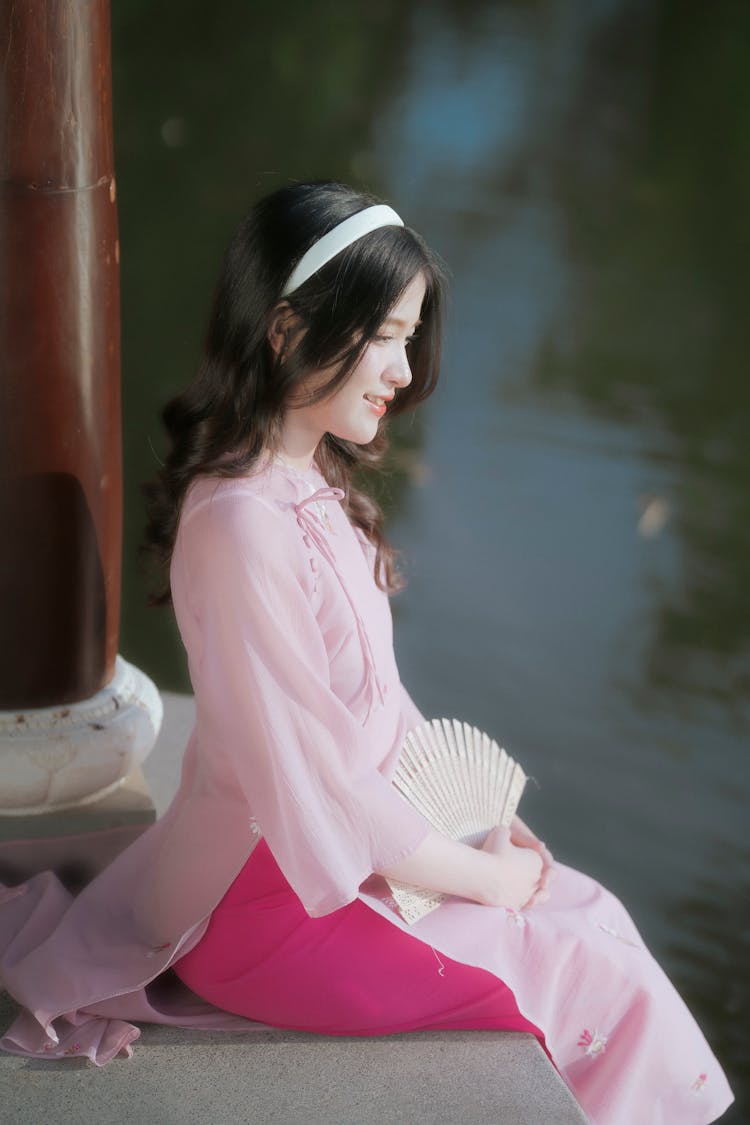 Young Woman In A Pink Ao Dai Dress Sitting By A Body Of Water