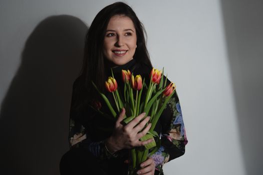 Smiling woman in a floral dress holds vibrant tulips, exuding warmth and joy in a studio setting.