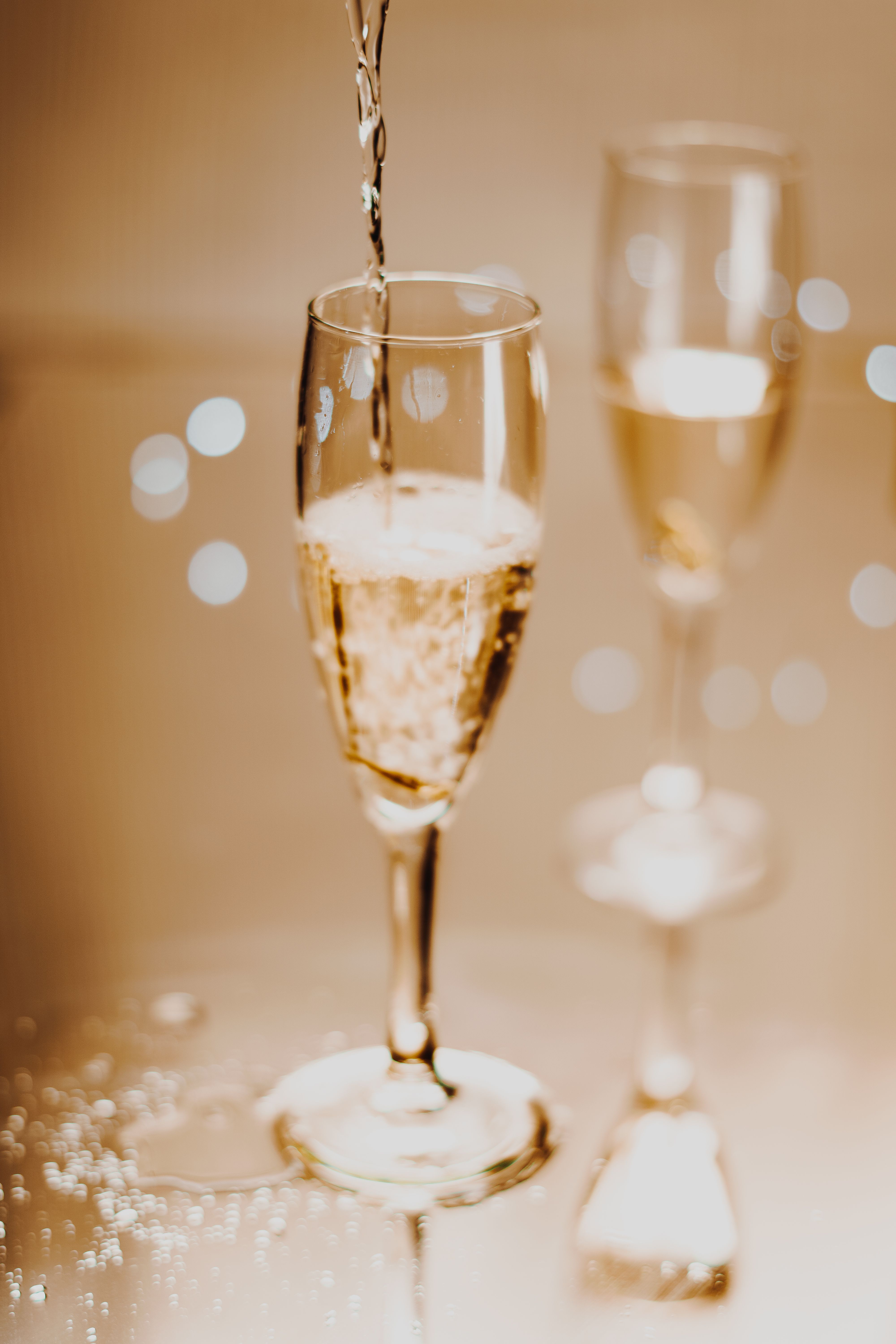 Two flutes of pale golden prosecco being poured at an angle, bubbles rising, with a green glass bottle blurred in the background.