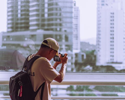 Man in Cap and with Backpack Taking Pictures in City