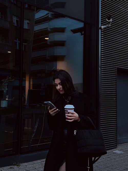 Young Woman Carrying a Smartphone and a Cup of Coffee Walking on the Sidewalk
