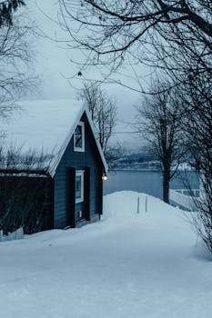 A serene winter scene of a snowy cabin by a frozen lake in Laksvatn, Norway.