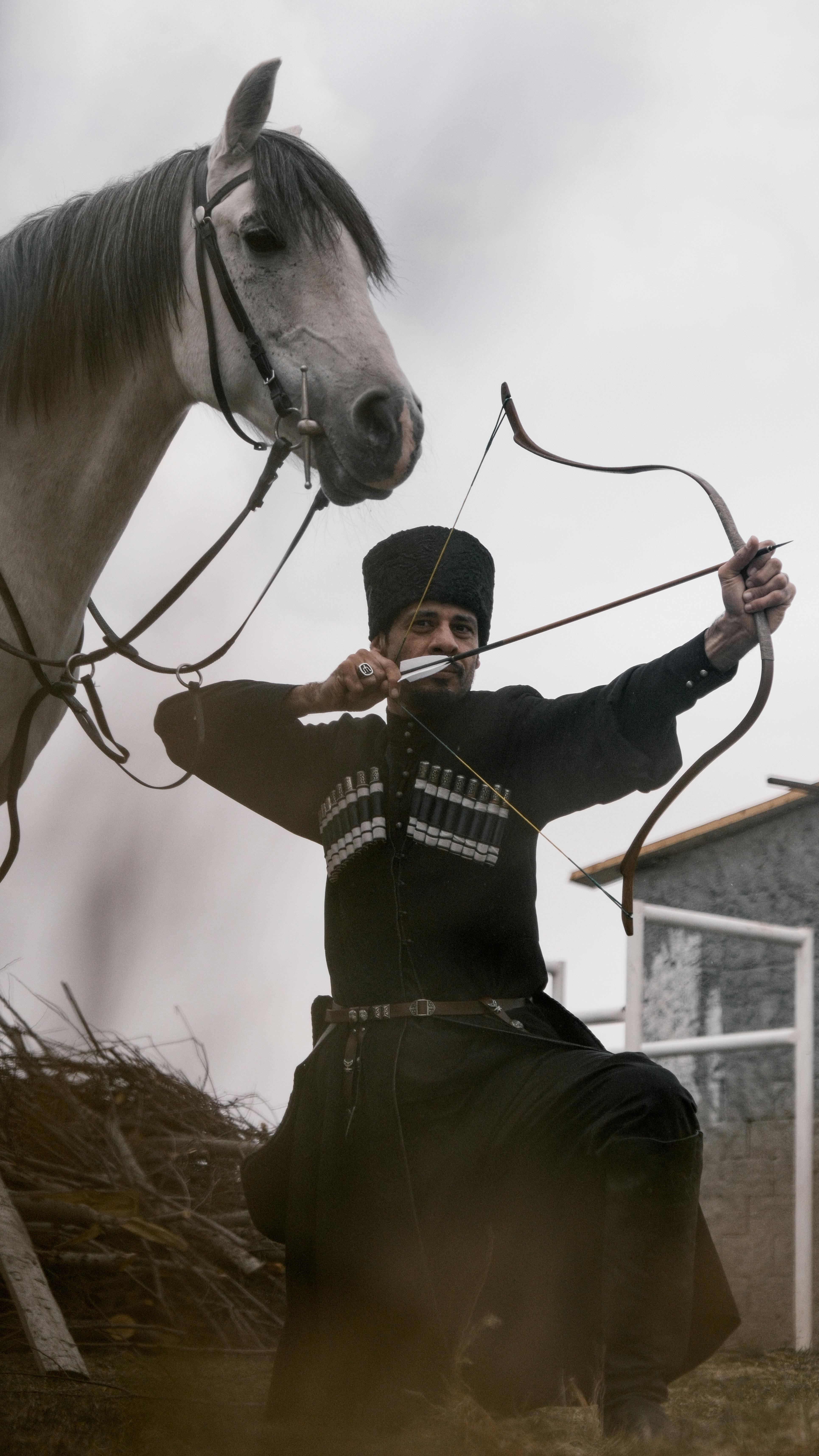 Man in traditional Georgian costume aiming a short bow beside a horse outdoors.