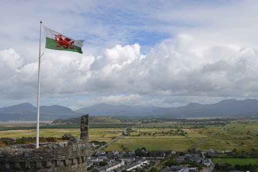 A Welsh flag waves over a scenic countryside landscape with mountains and a town visible.