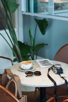 A warm café scene featuring a latte, vintage camera, book, and sunglasses on a white table.