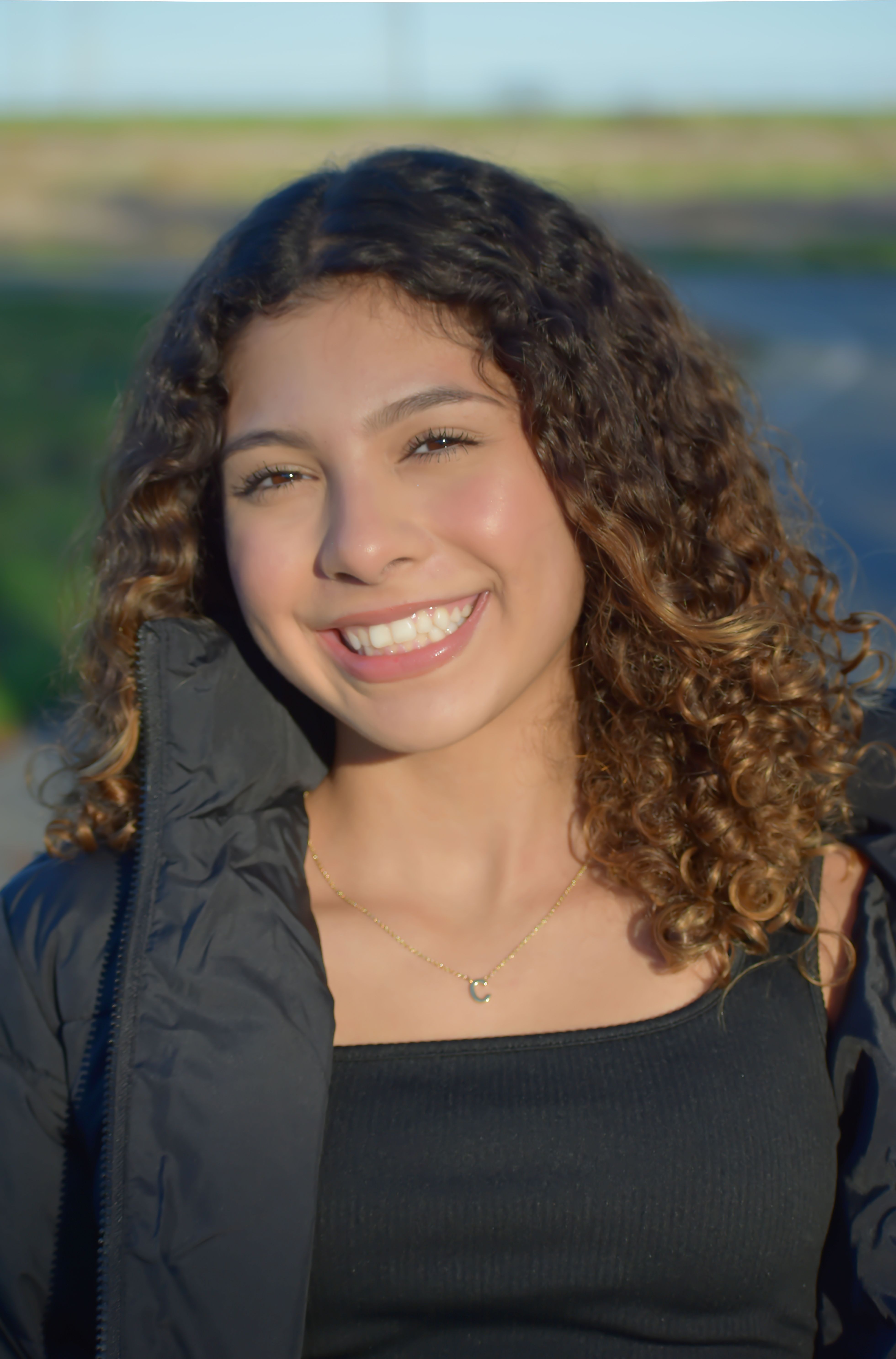 Cheerful woman with curly hair and necklace smiling outdoors during the day.