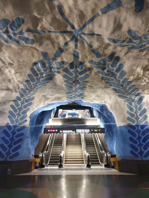 Vibrant blue and white artwork adorns Stockholm's metro station ceiling above an escalator.