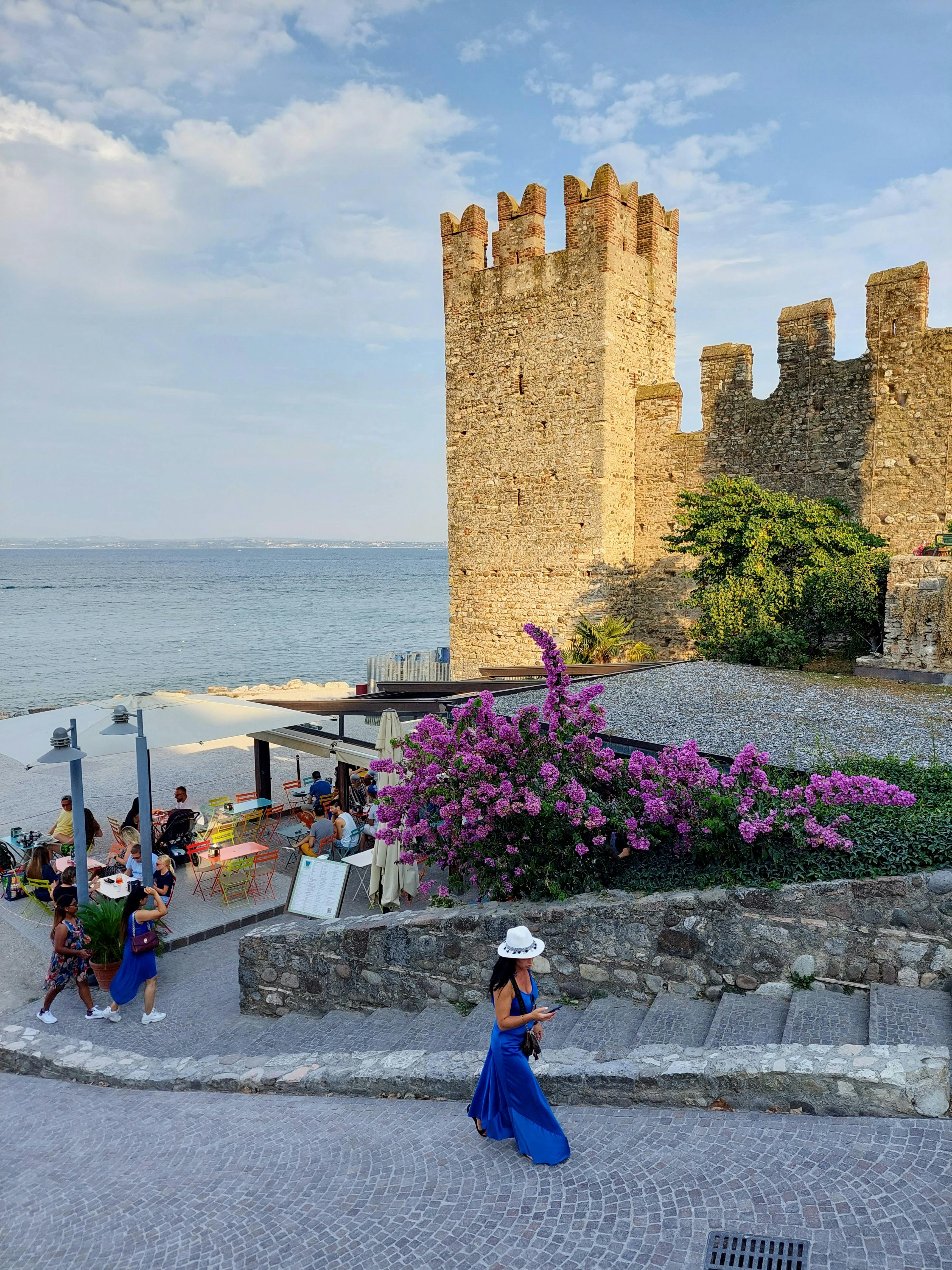 Woman in blue dress walking near Scaliger Castle in Sirmione with pink flowers and Lake Garda view.