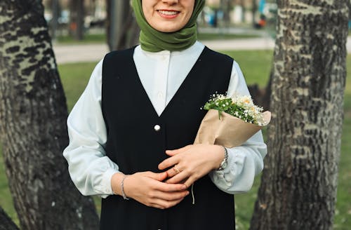 Free Smiling Woman in Vest and Shirt Standing with Flowers between Trees Stock Photo