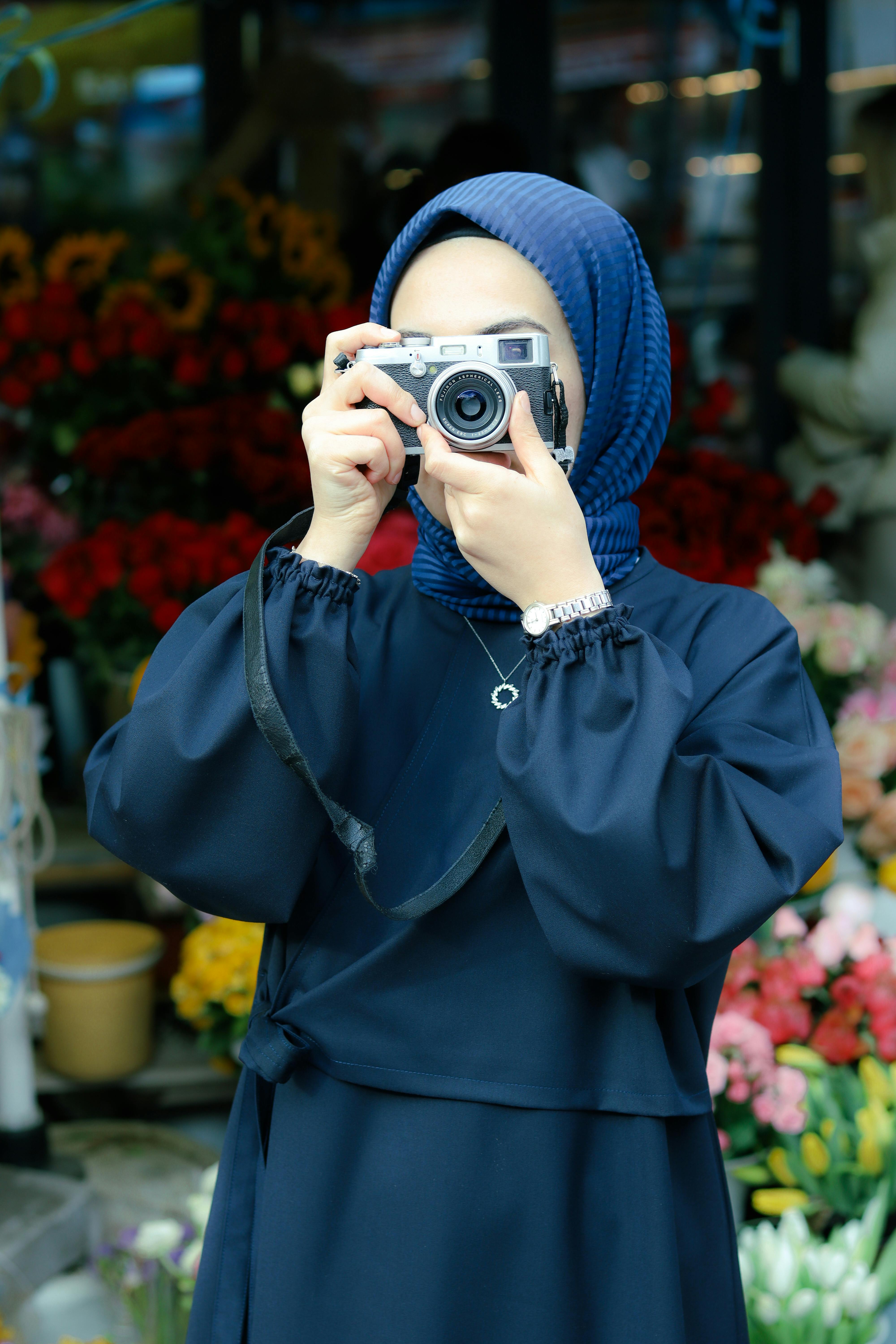 A woman wearing a hijab captures a photo with an analogue camera in a vibrant outdoor setting.