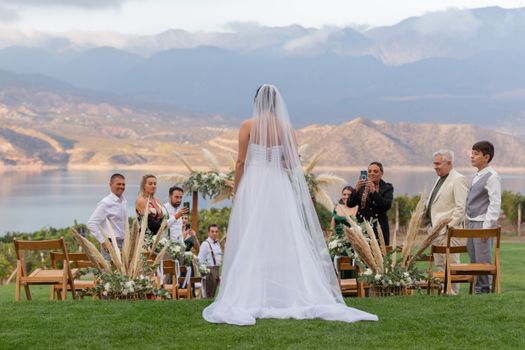 Bride in a wedding dress stands before guests at an outdoor ceremony overlooking a lake.