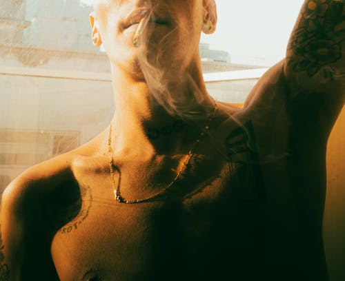 A man with tattoos on his chest smokes a cigarette