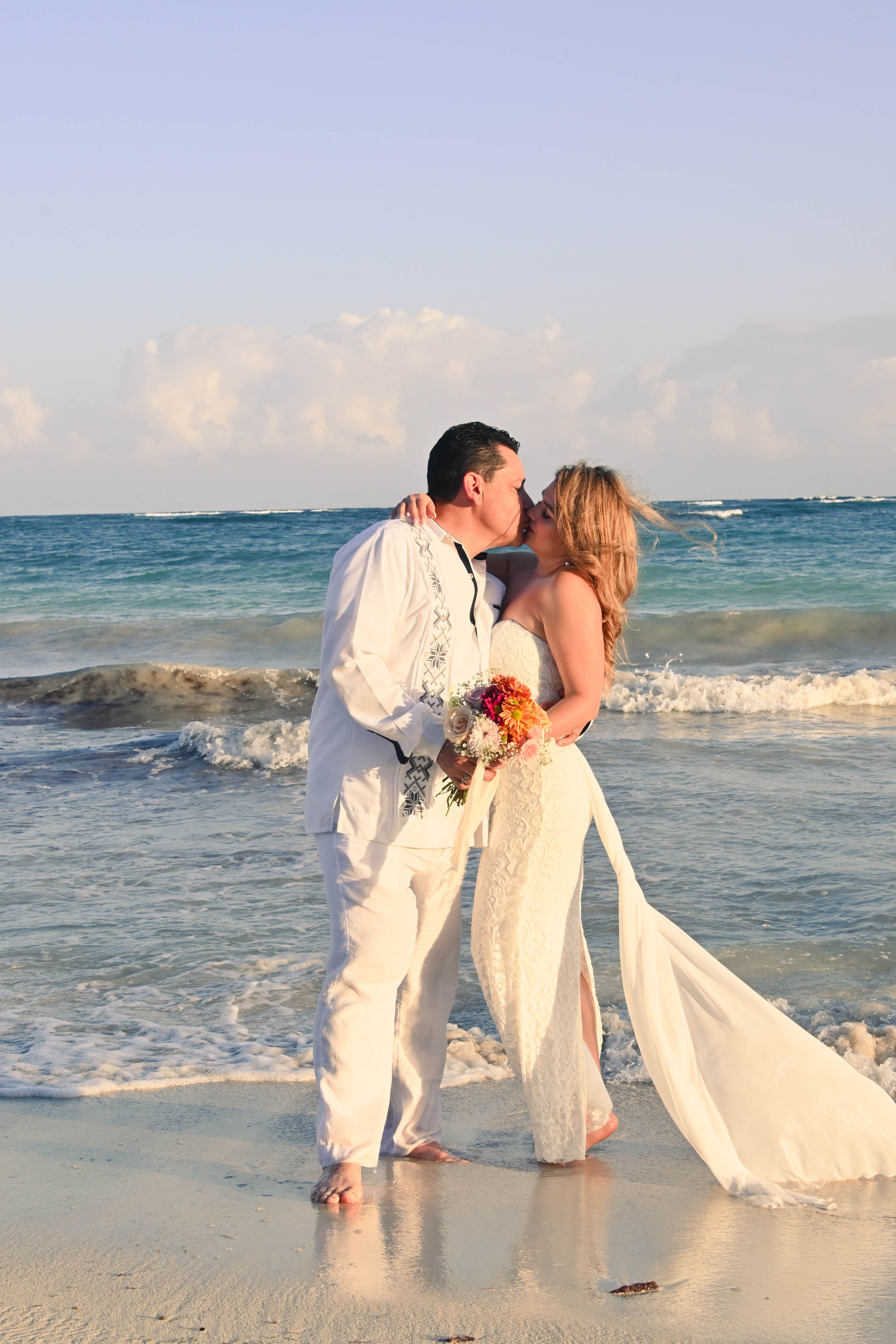 A couple shares a romantic kiss on the beach, framed by ocean waves and bright sunshine.