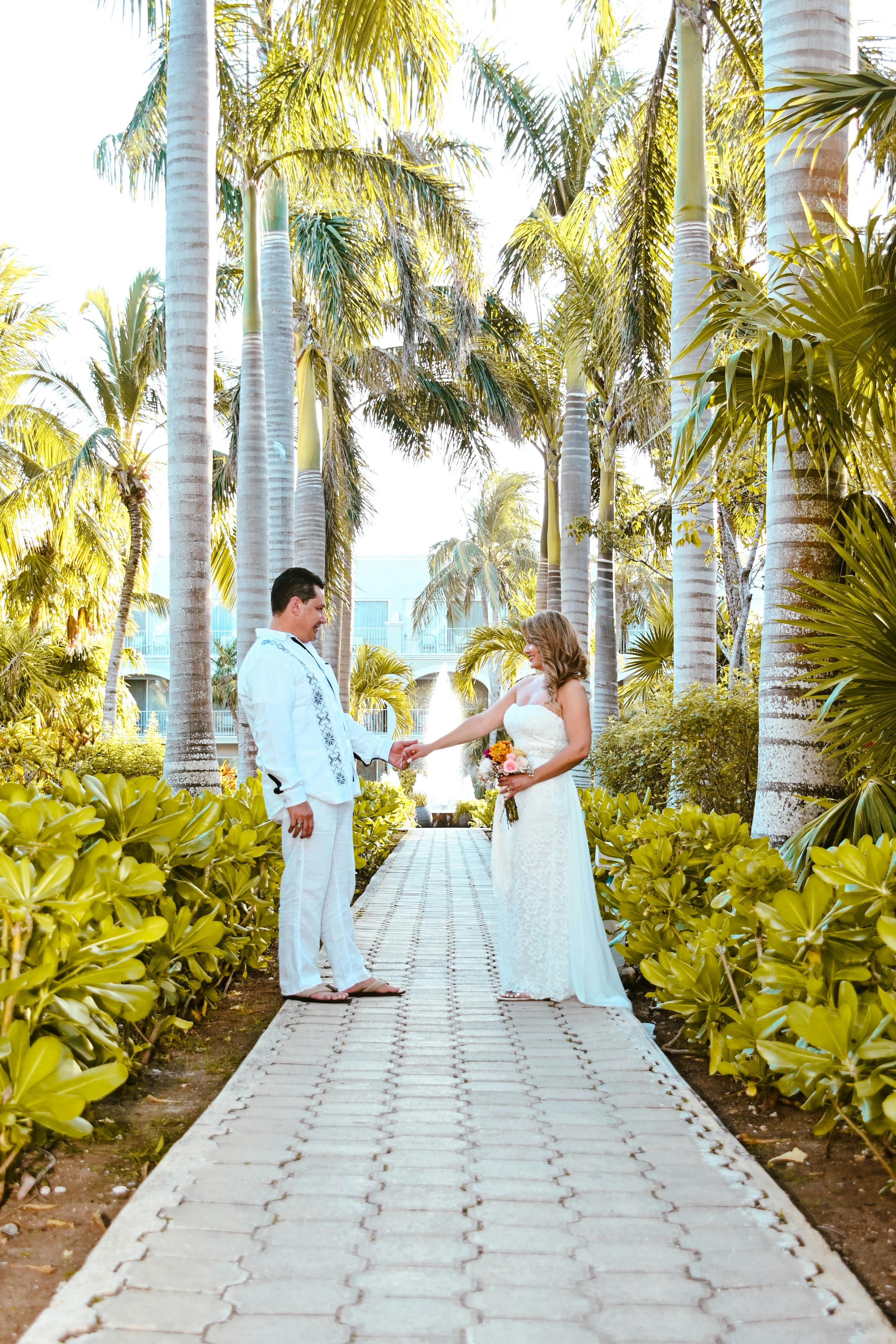 A bride and groom in a white dress and suit standing in a lush palm-lined pathway, exchanging vows.