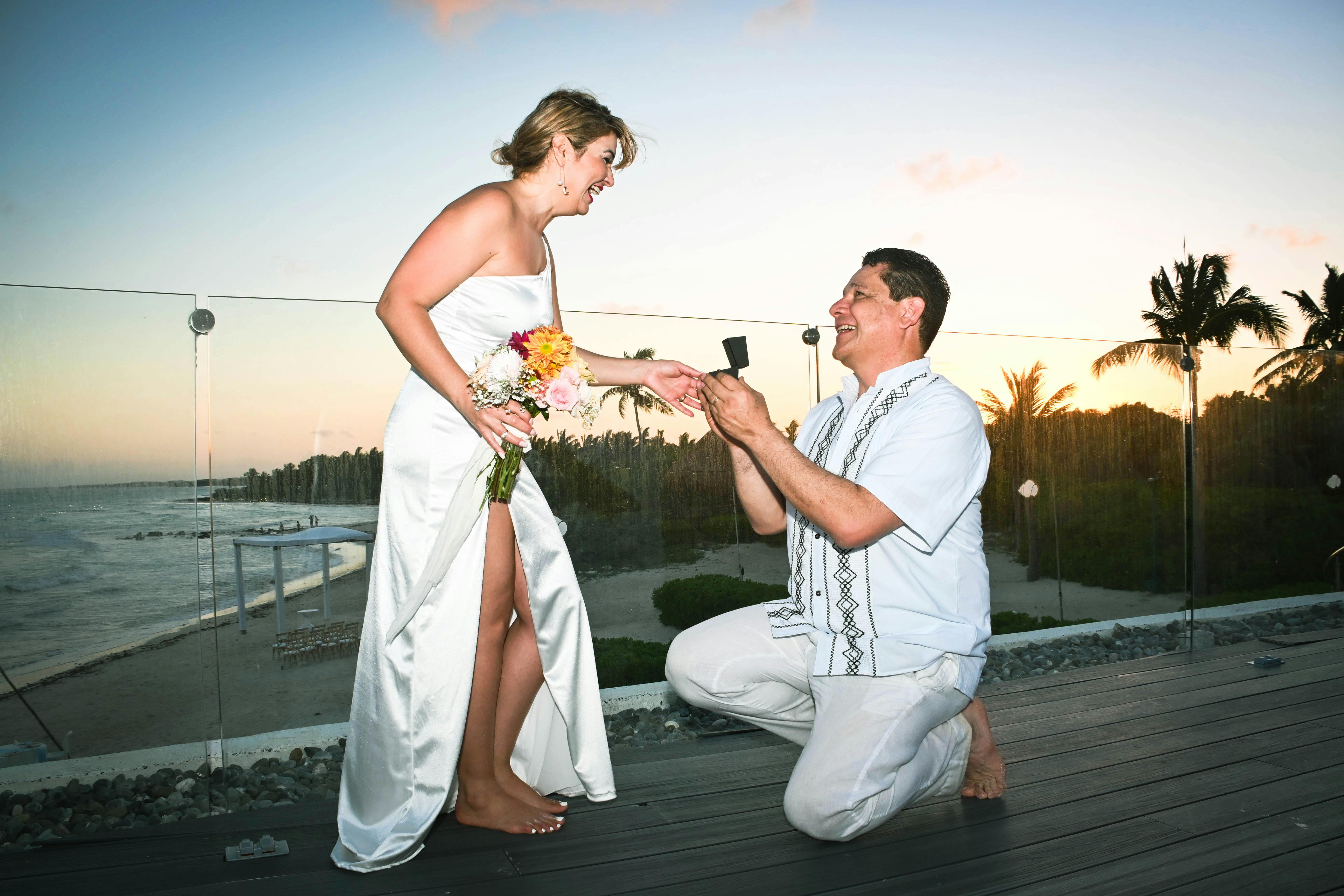 A joyful couple's beachfront proposal at sunset, with the man kneeling to present a ring.