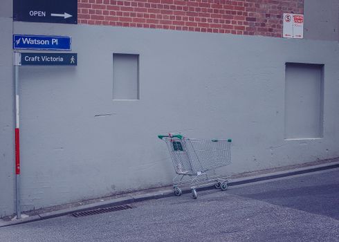 An empty shopping cart rests by a brick wall on a city street corner.