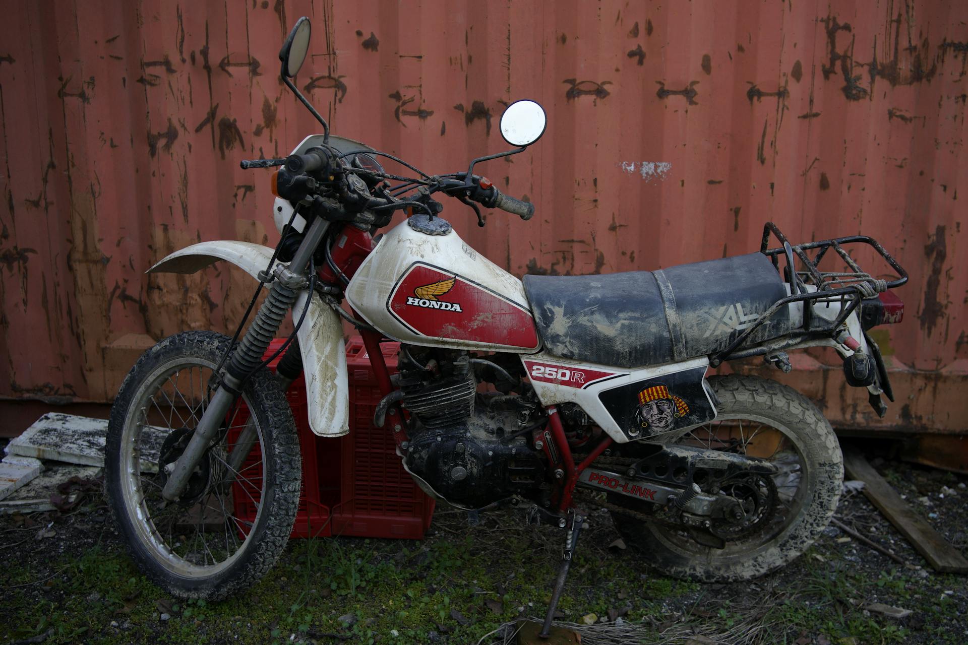 A dirt-covered Honda 250R motorbike by a rusty container in Faenza, Italy.