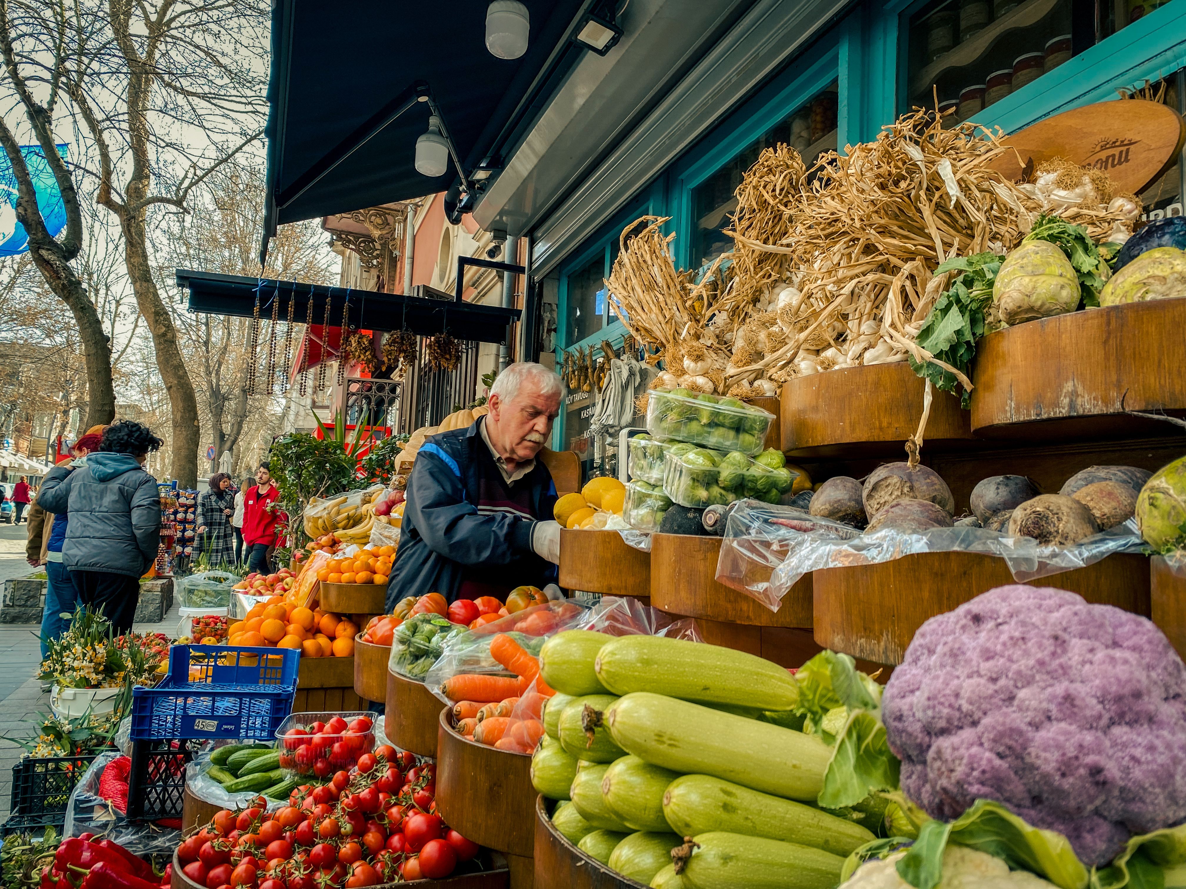A colorful, bustling street market in a Balkan city with fresh produce, locals, and vintage cars