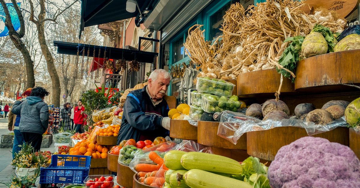 A colorful, bustling street market in a Balkan city with fresh produce, locals, and vintage cars
