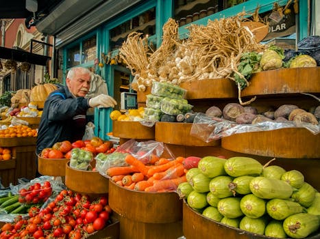 An elderly man selects vegetables at a vibrant outdoor urban market stand filled with fresh produce.