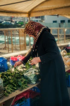 A woman selects fresh herbs from a market stall, showcasing local produce in an outdoor setting.
