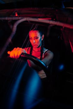 A woman with braided hair drives at night, illuminated by vivid red and blue lights.