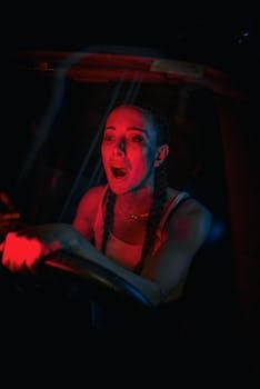 A woman with braids captured in the red-lit interior of a car, driving at night.
