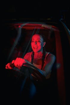 A woman driving a car at night, illuminated by dramatic red and blue light, expressing intense emotion.
