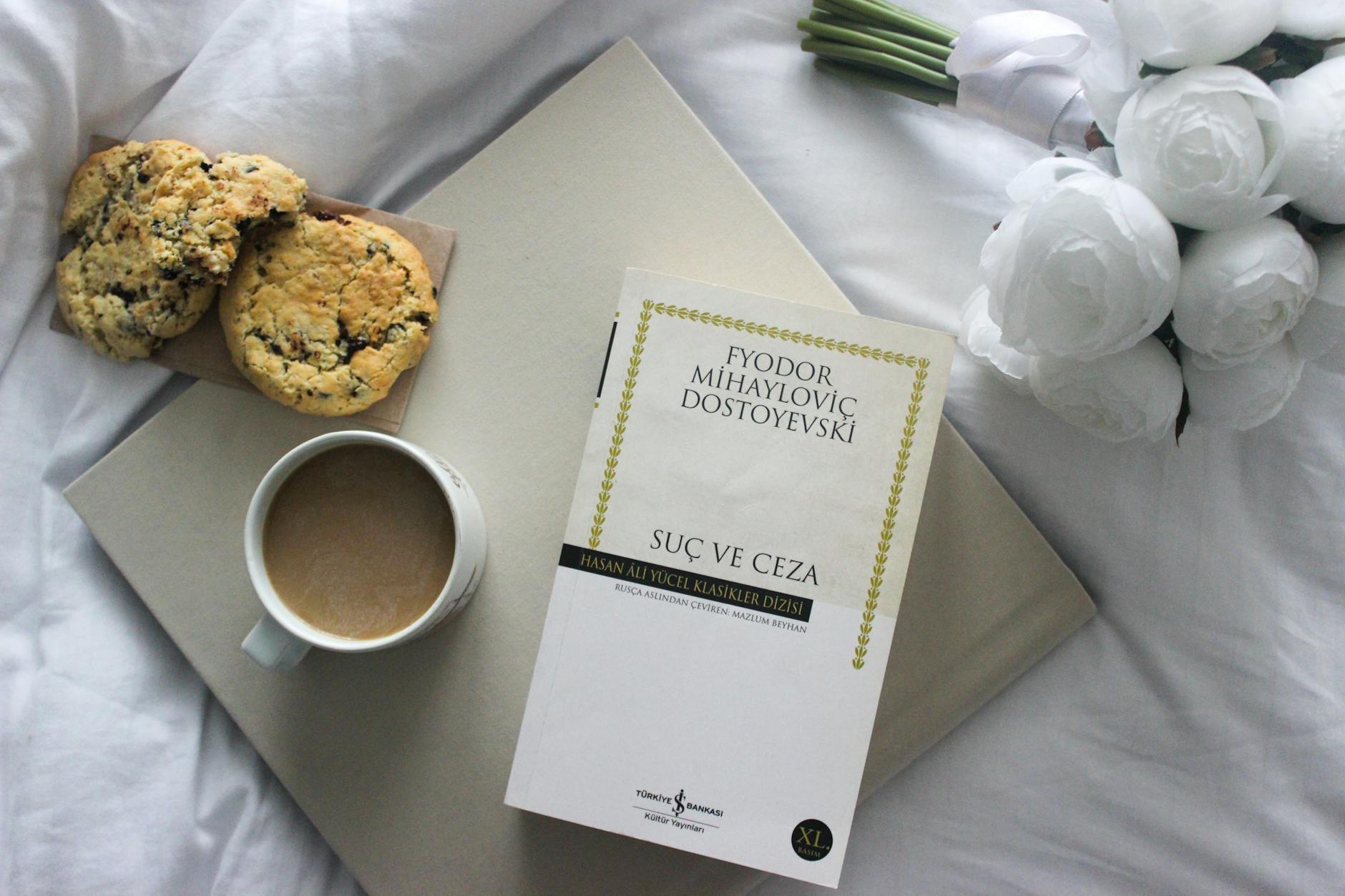 Top view of a book, coffee, and cookies on a bed with white roses.