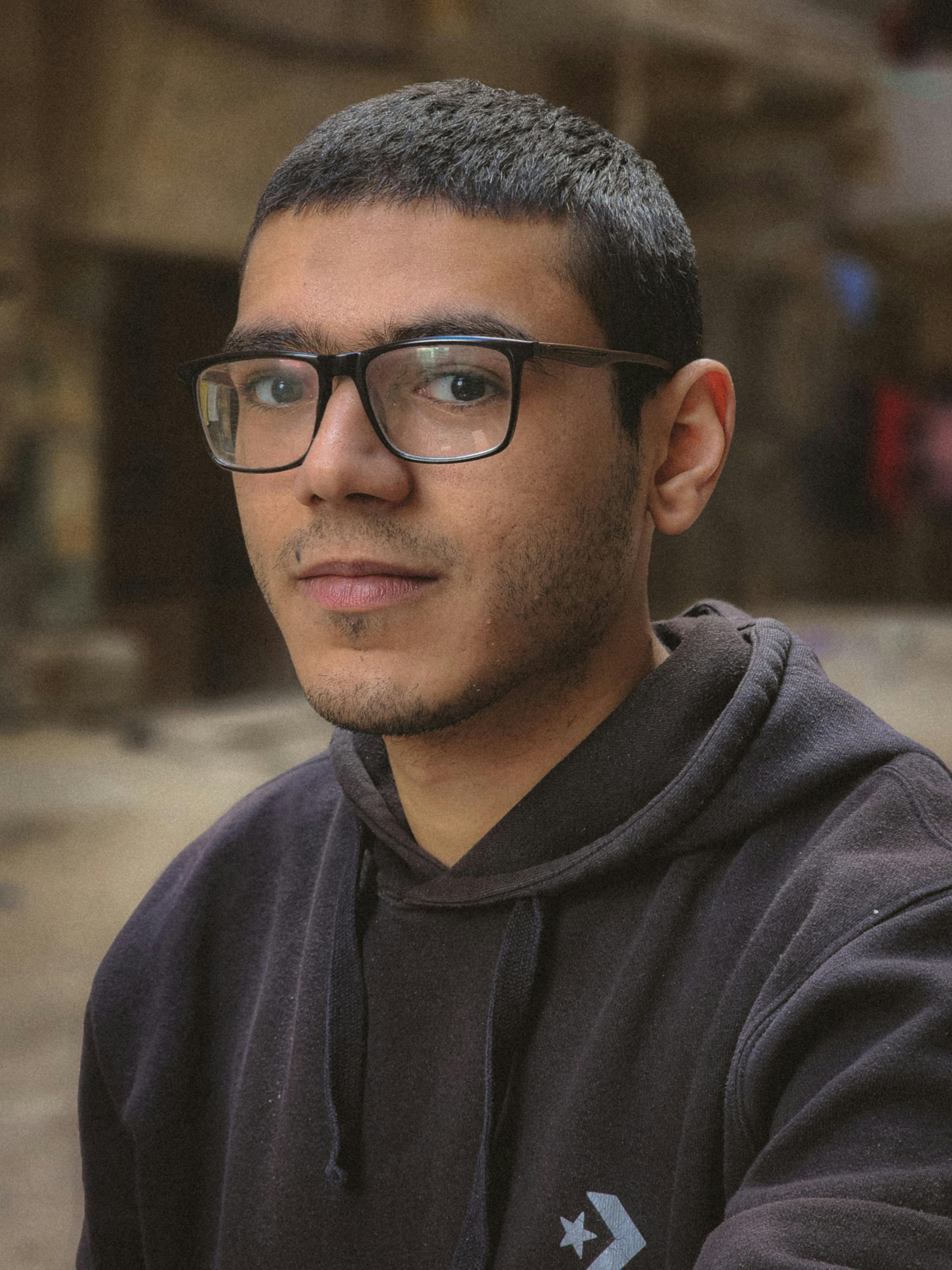 Close-up portrait of a young man with glasses in urban setting, wearing a hoodie.