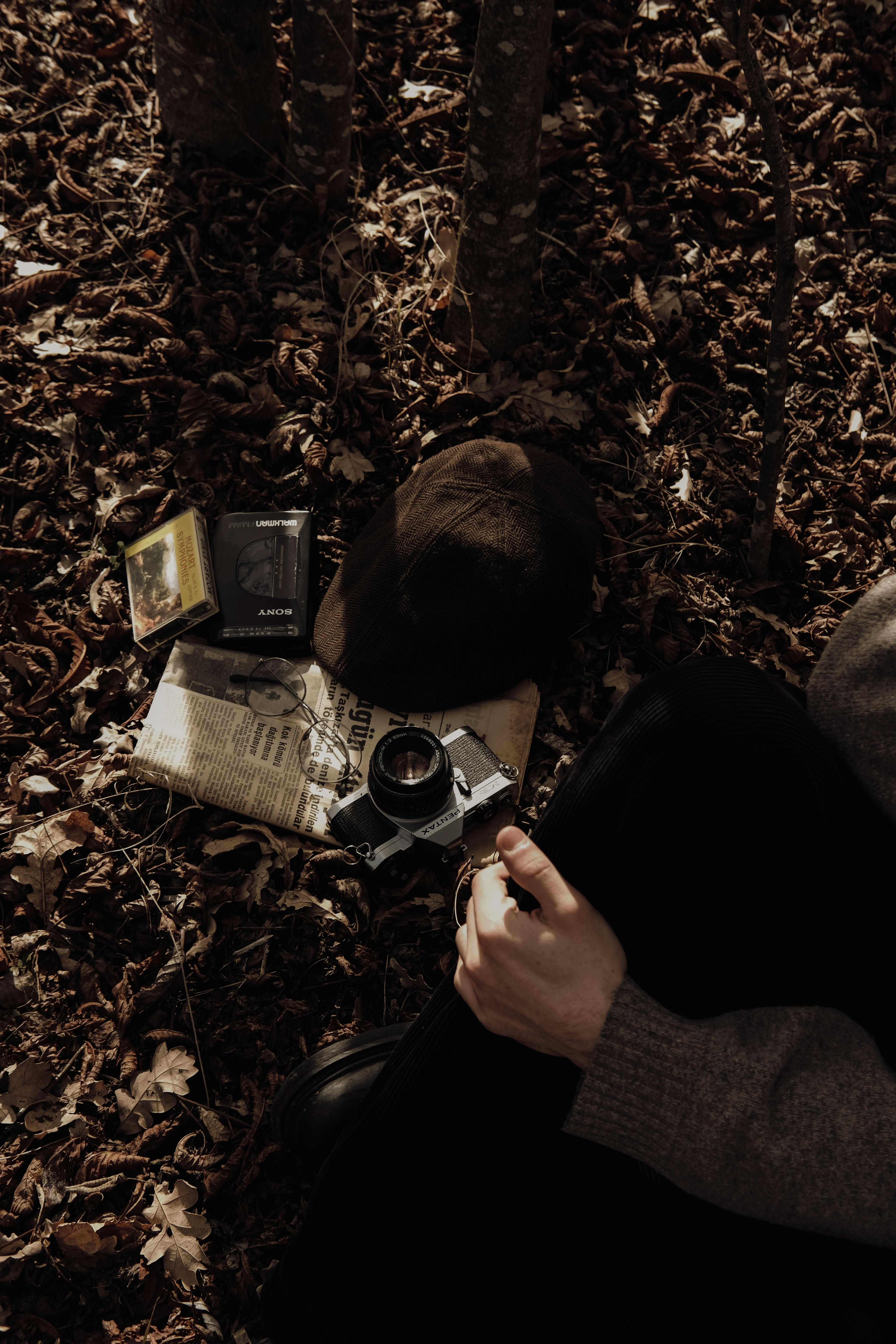 Dark, moody autumn setting with vintage camera, cap, and newspaper on forest ground.