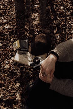 A vintage camera rests among fallen leaves beside a person's hands in an autumn forest setting.
