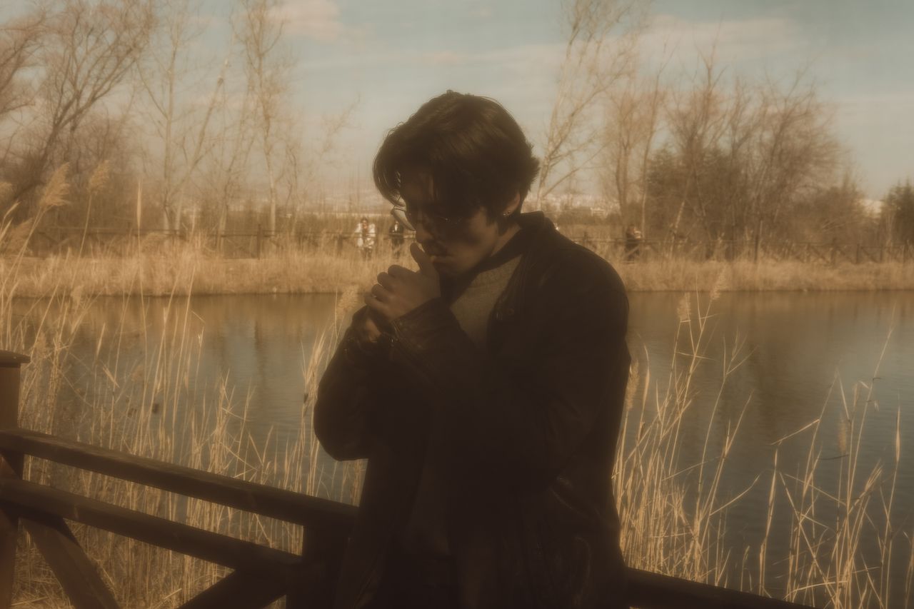 a young man lights a cigarette by a lake surrounded by autumn trees in