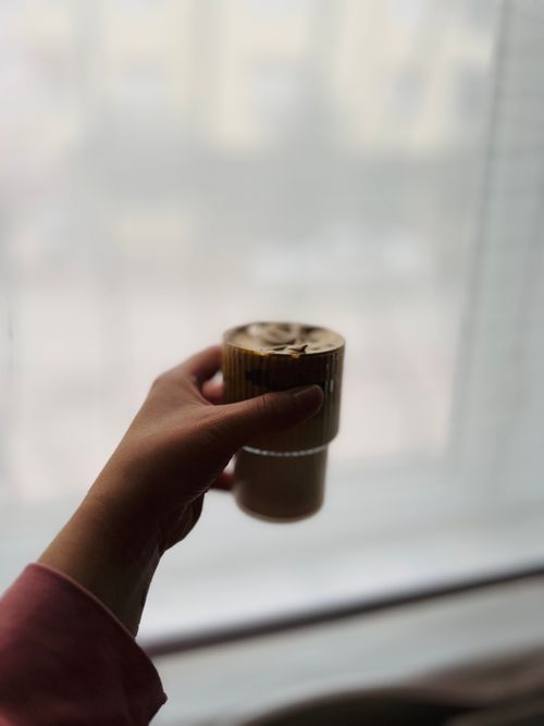 Free Hand holding a cup of coffee with foam in front of a blurred window background. Stock Photo