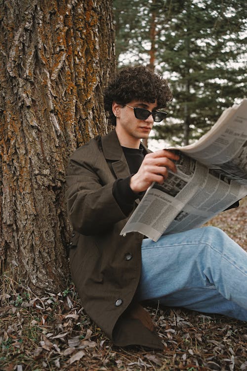 Man in Sunglasses and Jacket Sitting by Tree and Reading Newspaper
