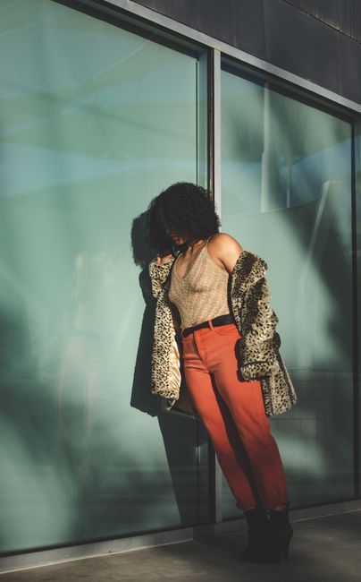 Fashionable woman in leopard print coat leaning on modern glass building wall, showcasing bold urban style.