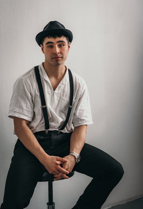 A man in suspenders and a hat sitting on a stool